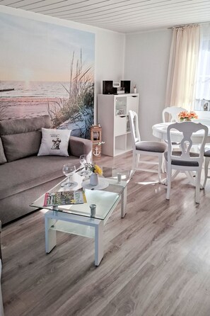 Een moderne woonkamer met een glazen salontafel, een bank, een eettafel met stoelen, een boekenkast en een groot schilderij van een strandgezicht.
