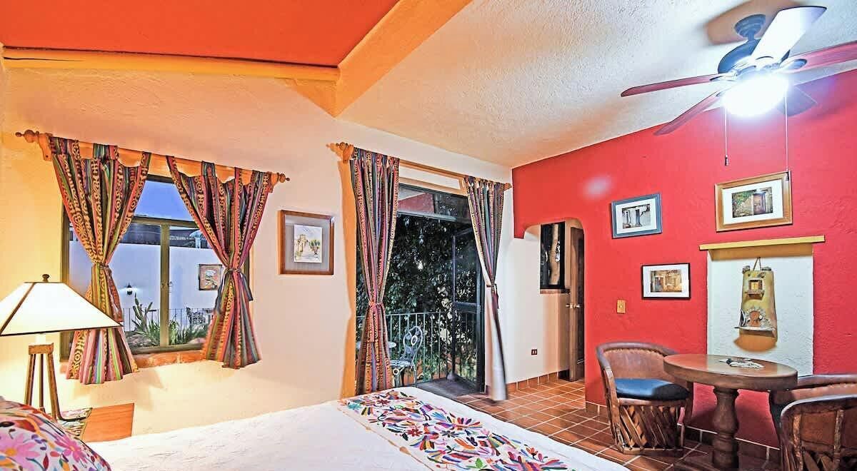 Une chambre d’hôtel avec un lit, une table et des chaises, un ventilateur de plafond et un balcon avec des rideaux.