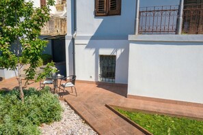 A courtyard with a patio, a small table with chairs, a potted plant, and a window with a metal grill. - Apartment Akidado - Garden & Sea View (Pula)