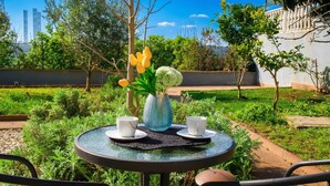 A round glass table with two white cups and a vase with flowers, surrounded by greenery and a clear sky.
