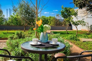 A round glass table with two white cups and a vase with flowers, surrounded by greenery and a clear sky.
