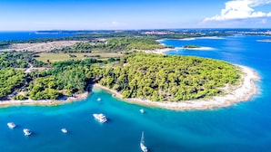 An aerial view of a coastal area with a small island surrounded by clear blue waters, several boats, and a distant cityscape.