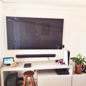 Televisão de tela plana na parede, escrivaninha com computador, um controle de videogame rosa e duas plantas em vasos.