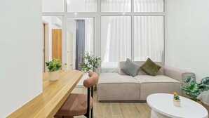 A modern living room with a wooden dining table, a sofa with cushions, a white coffee table, and large windows with sheer curtains.