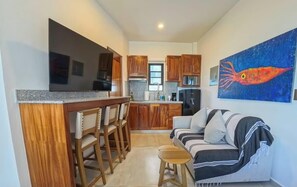 Eine kompakte Küche mit Holzausstattung, ein Essbereich mit Barhocker und ein Wohnzimmer mit Sofa und einem Gemälde.