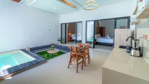 A modern hotel room with a large bed, a dining area with a table and chairs, a kitchen area with a coffee maker, and a small outdoor pool area. - Ghania Villas Melasti (Ungasan)