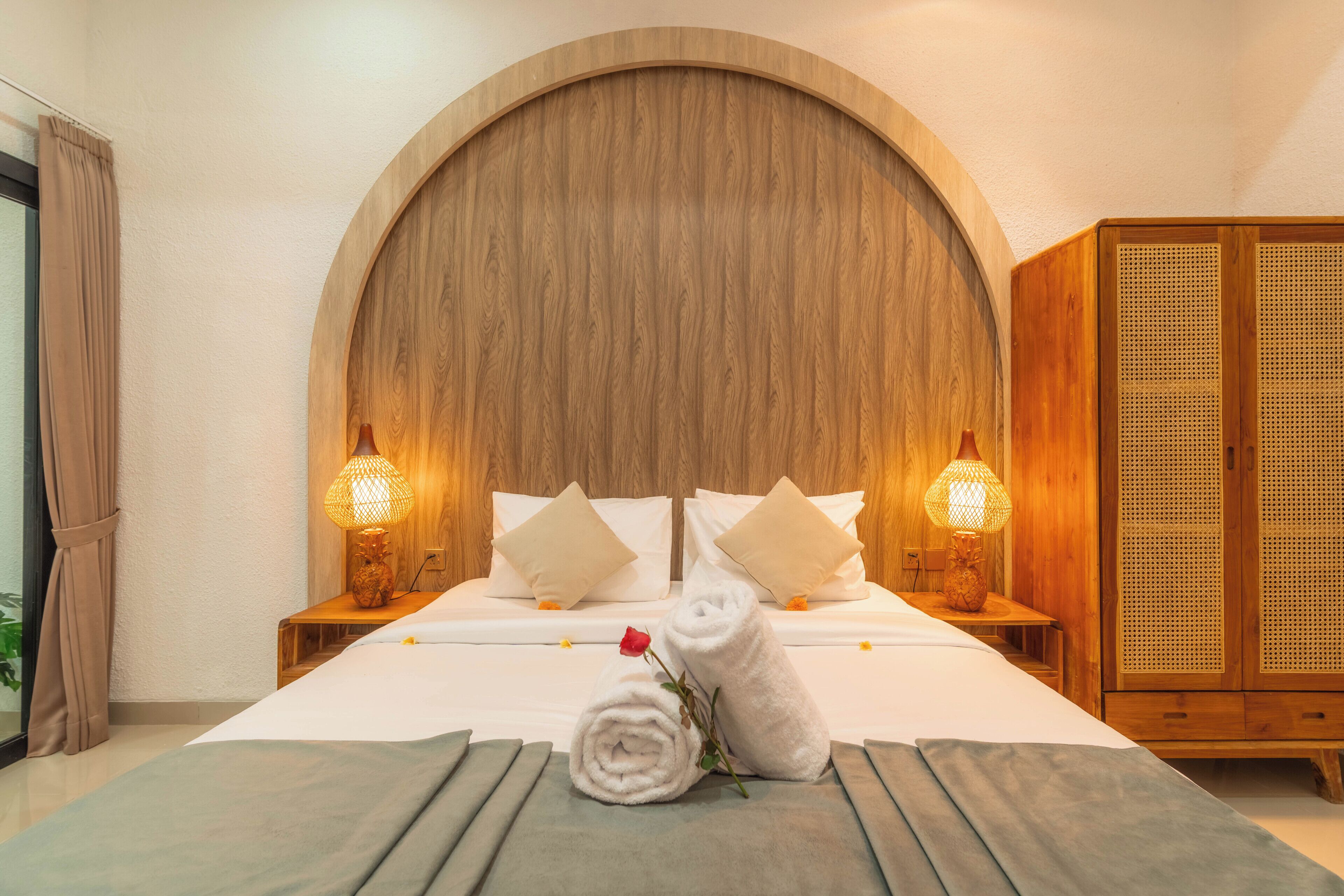 Habitación de hotel con una cama grande, dos lámparas de noche, un cabecero de madera, un armario y una toalla enrollada con una rosa en la cama.