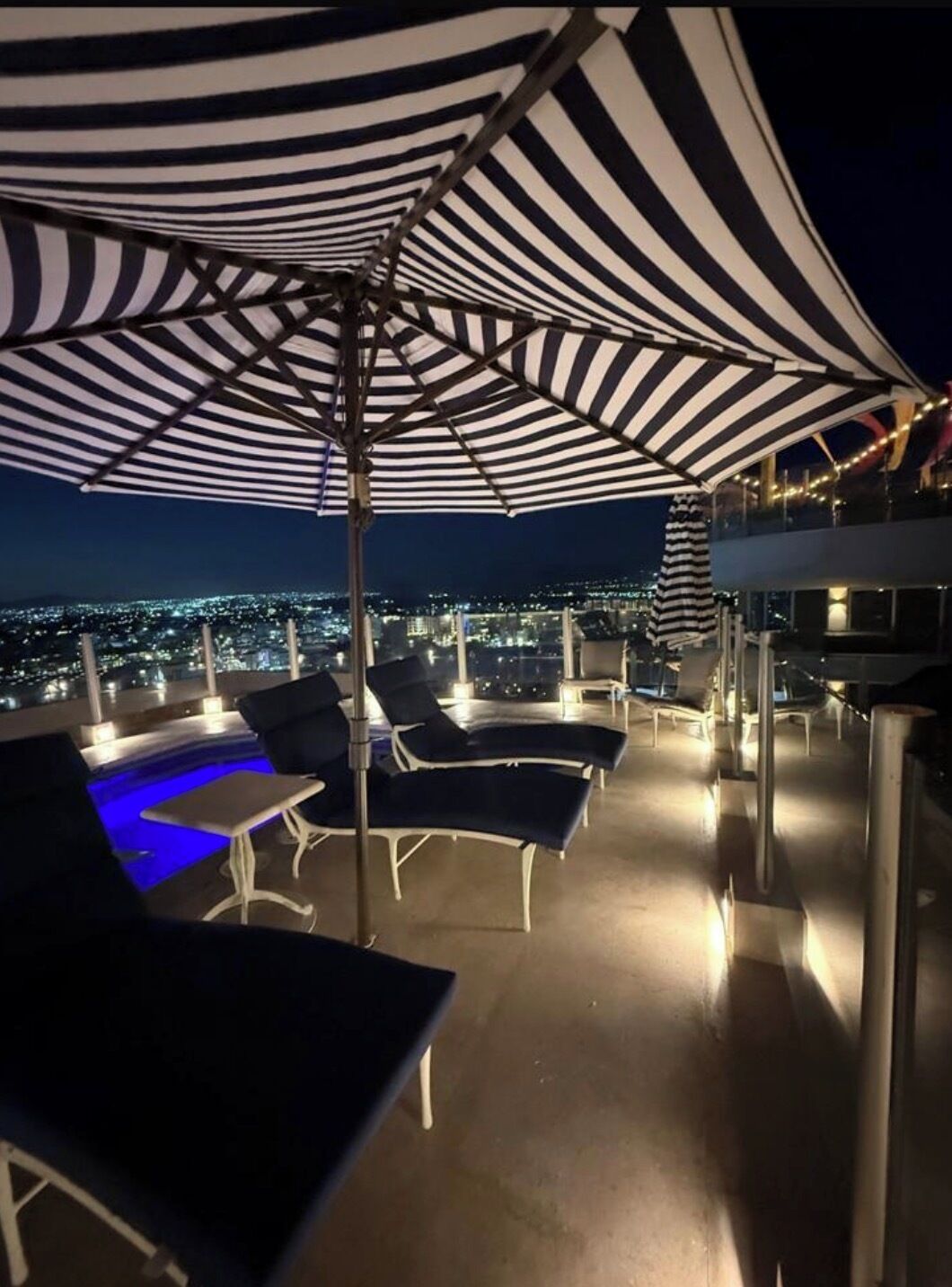 Eine Dachterrasse mit gestreiften Sonnenschirmen, Liegestühlen und Blick auf die Stadt bei Nacht.