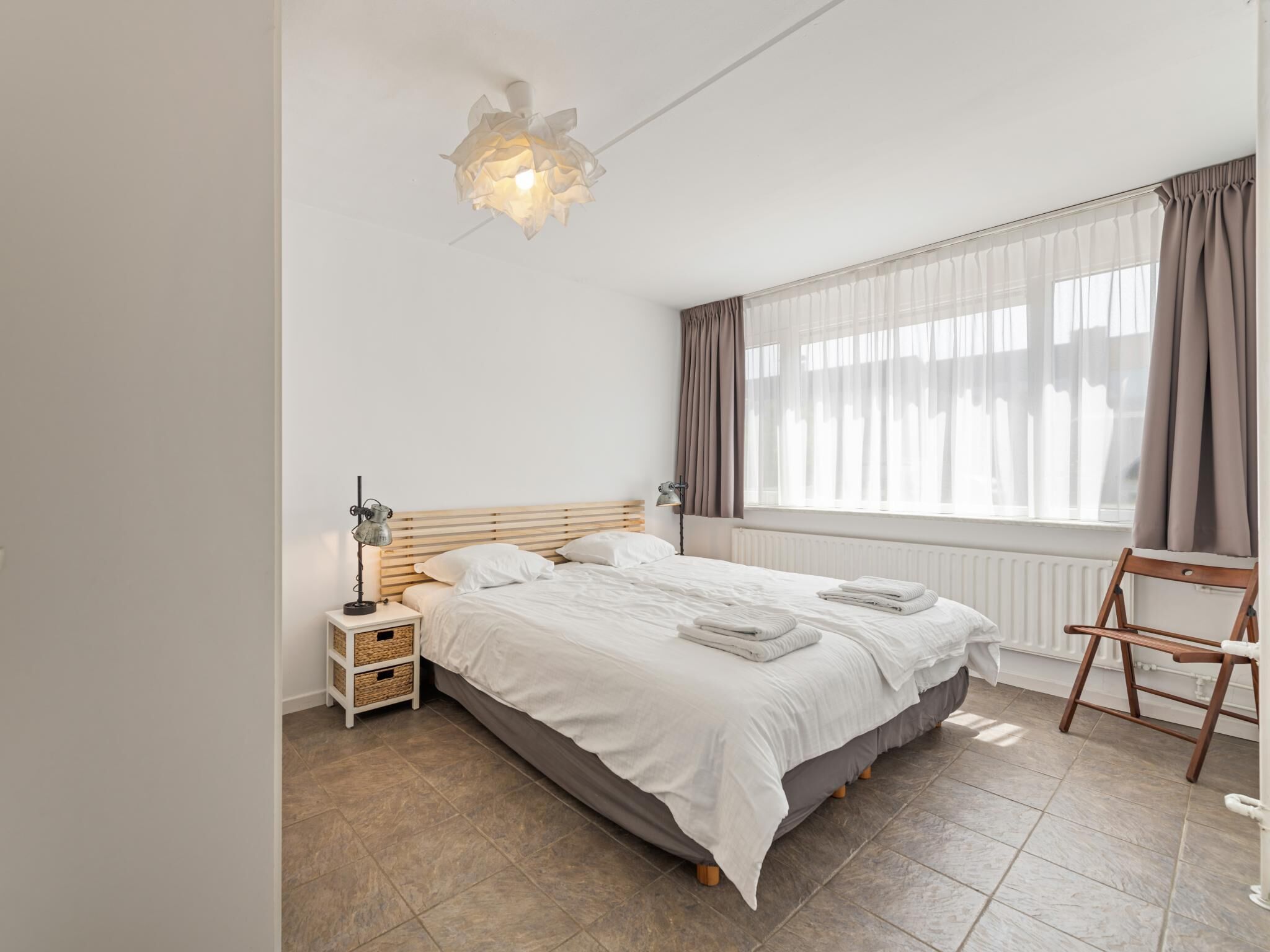 A bedroom with a bed, bedside tables, a lamp, a chair, a window with curtains, and a ceiling light fixture.