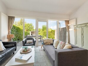 A spacious living room with a sofa, armchair, and coffee table, large windows, and a view of a garden. - Nice beach house directly by the sea with garden (Nieuwvliet)