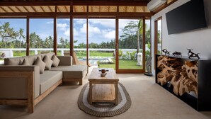 A spacious living area with a large window, a sofa, a coffee table, and a wall-mounted TV. - Uma Dawa Residence (Ubud)