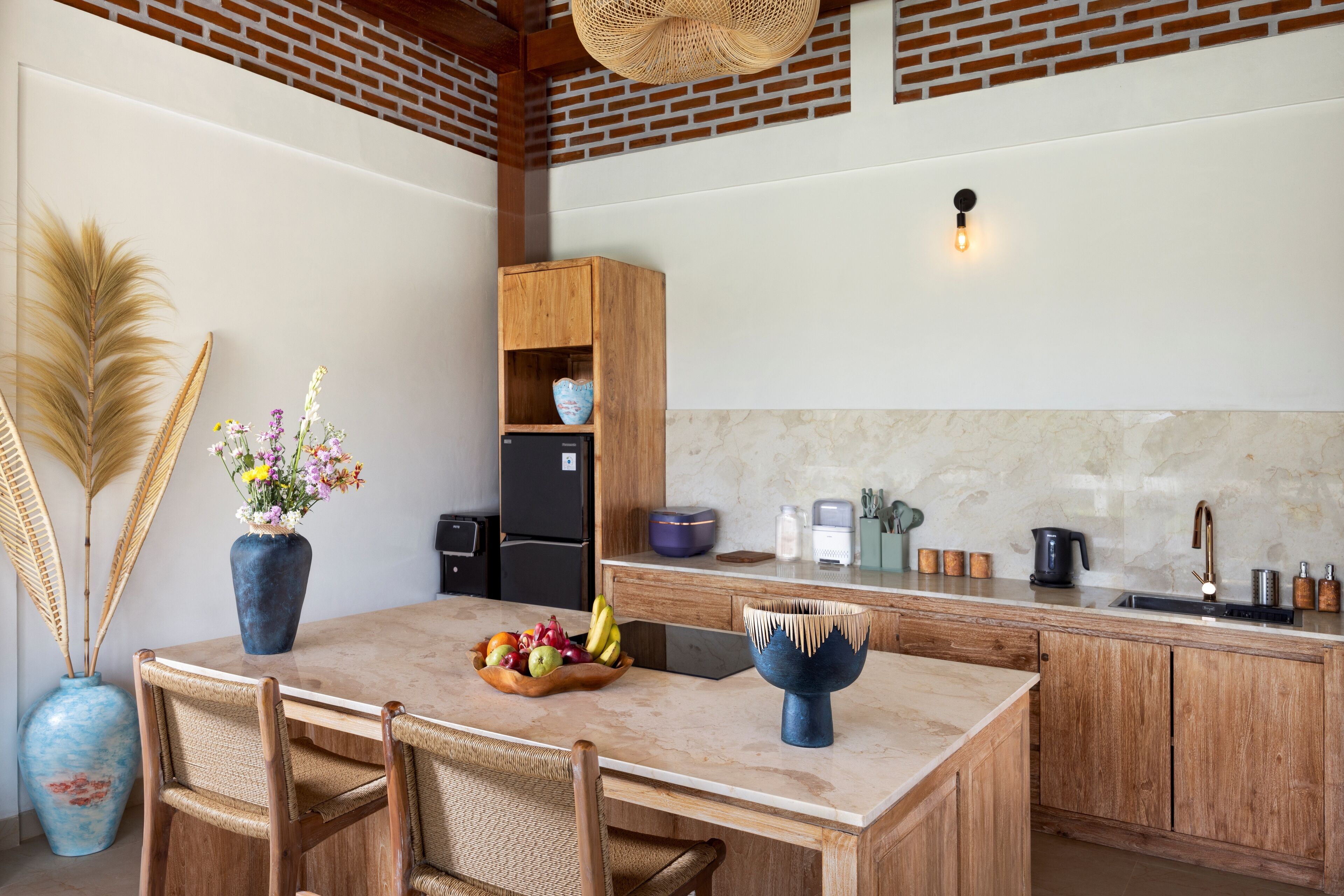 A kitchen with a wooden table, chairs, a vase with flowers, a fruit bowl, a blue bowl, a brick wall, a hanging light, and a black refrigerator.