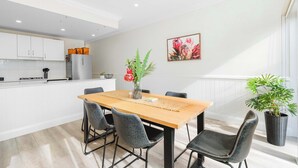 A modern kitchen with white cabinets, a central island, and a dining area with a wooden table and chairs. - Travers Townhouse - Close to CBD, Gated Community (Wagga Wagga)