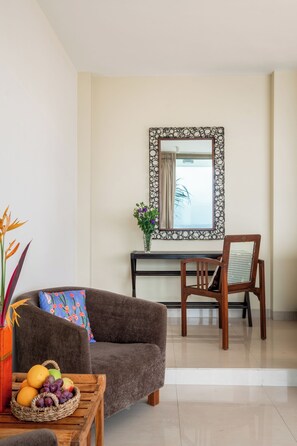 A living room with a sofa, a wooden chair, a small table with a fruit basket, a decorative mirror, and a potted plant. - Staymaster Edge Water- Sea View 4BR (Nerul)