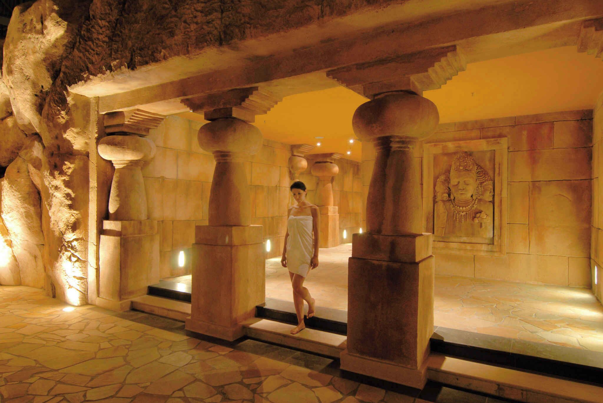 A stone corridor with columns, a person walking, and a statue on the wall.