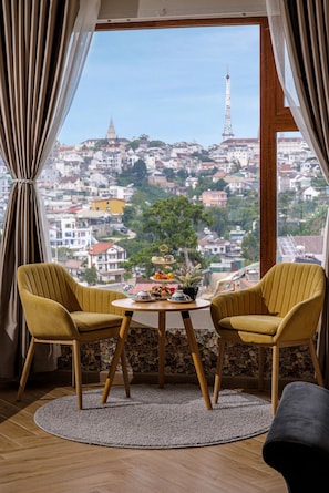 A room with two yellow chairs, a small table with a plant, and a view of a cityscape through a window. - Lacle- BB's Garden (Da Lat)