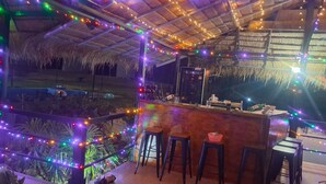Een bar met rieten dak, houten vloer en kleurrijke lichtslingers.