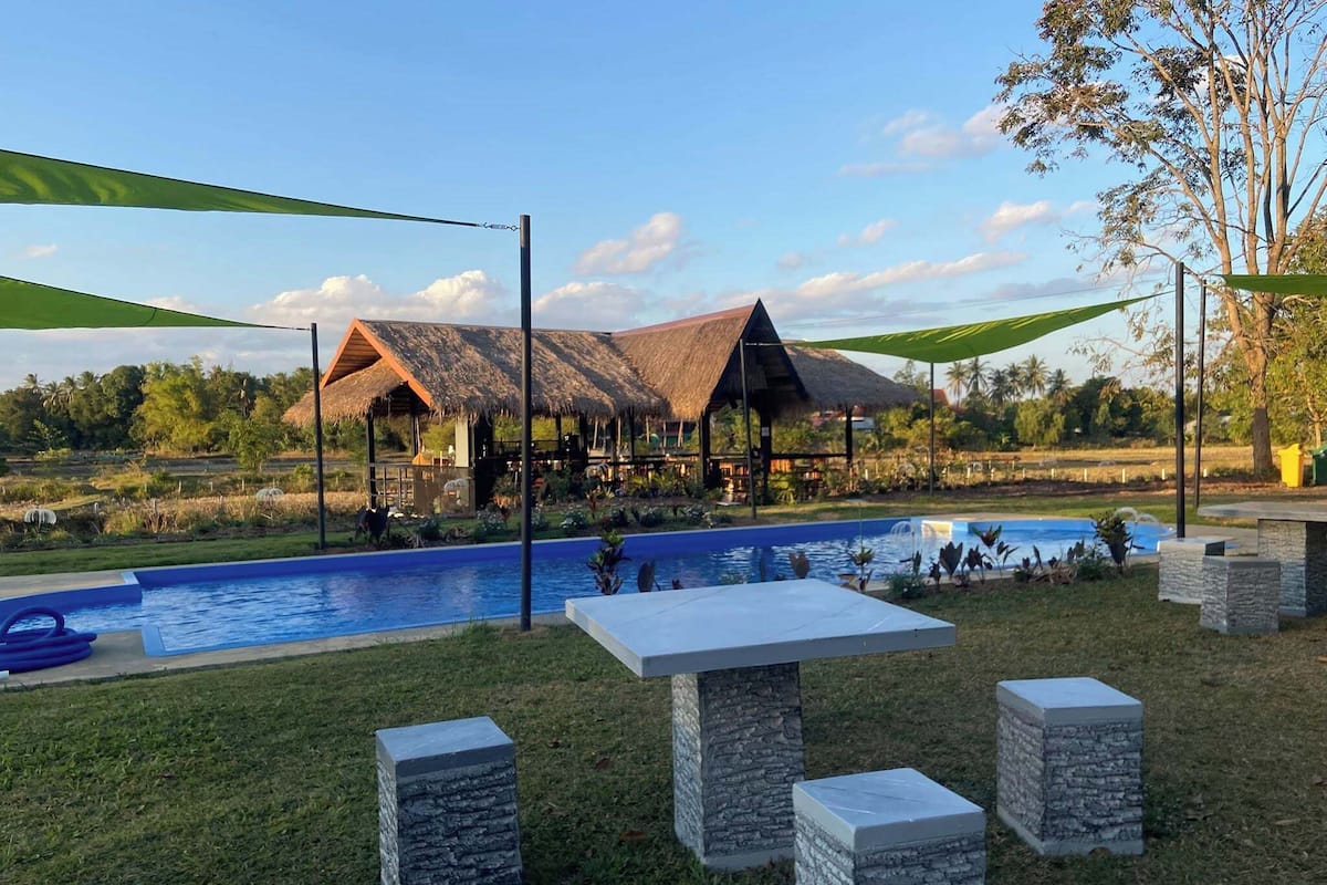 A pool area with a thatched-roof structure, stone pillars, and a clear sky.