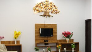 Ein modernes Hotelzimmer mit Flachbildfernseher, Deckenventilator, einer großen dekorativen Baumleuchte, einer Couch, einem Bett und einem kleinen Holztisch mit Blumenvasen.