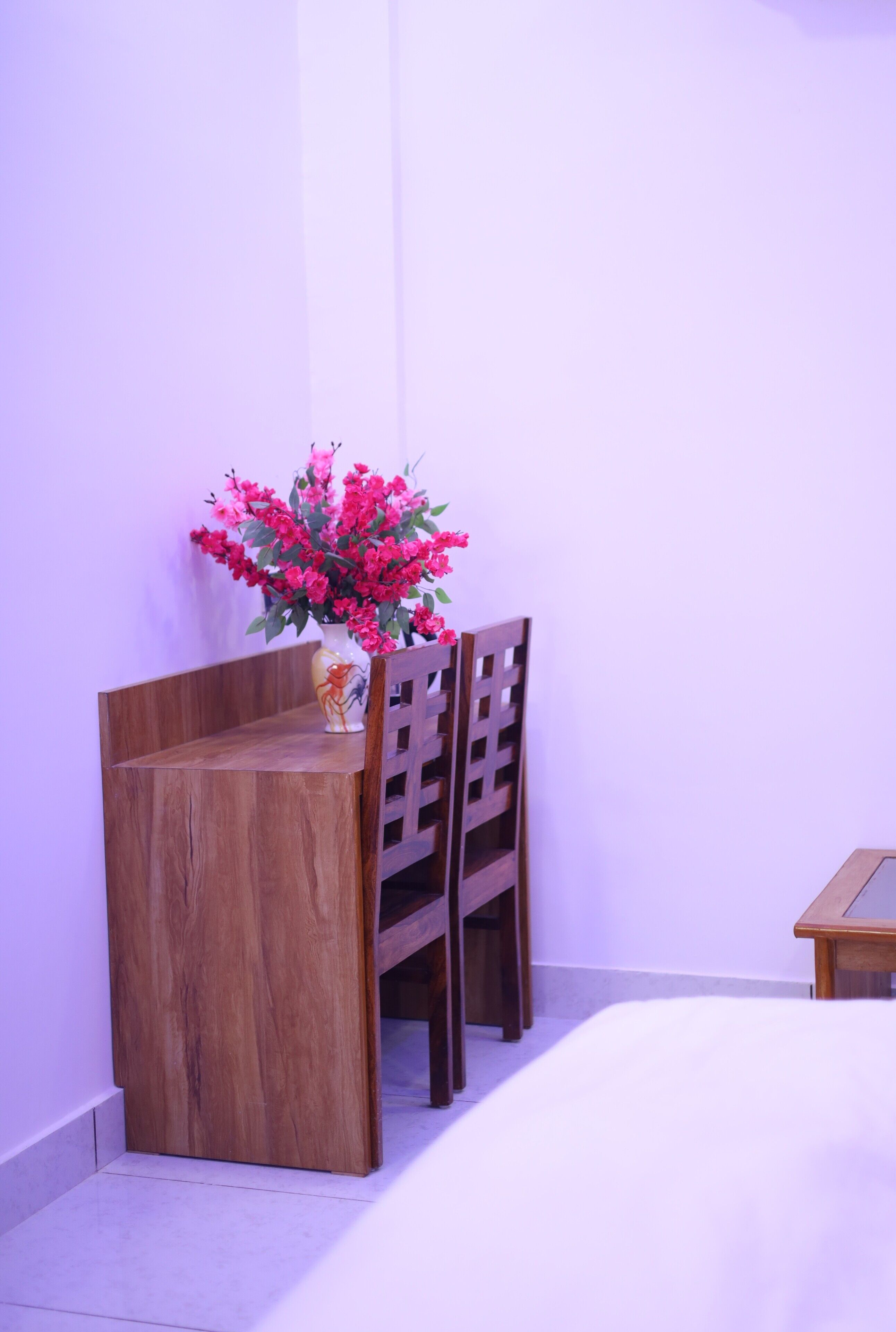 A wooden desk with a vase of flowers, a chair, and a small table against a plain wall.