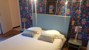 A bed with white linens, two pillows, a headboard, a bedside table with a lamp, and a wall with floral wallpaper. - Bed and breakfast La Villa Stella in Arcachon with swimming pool (La Teste-de-Buch)