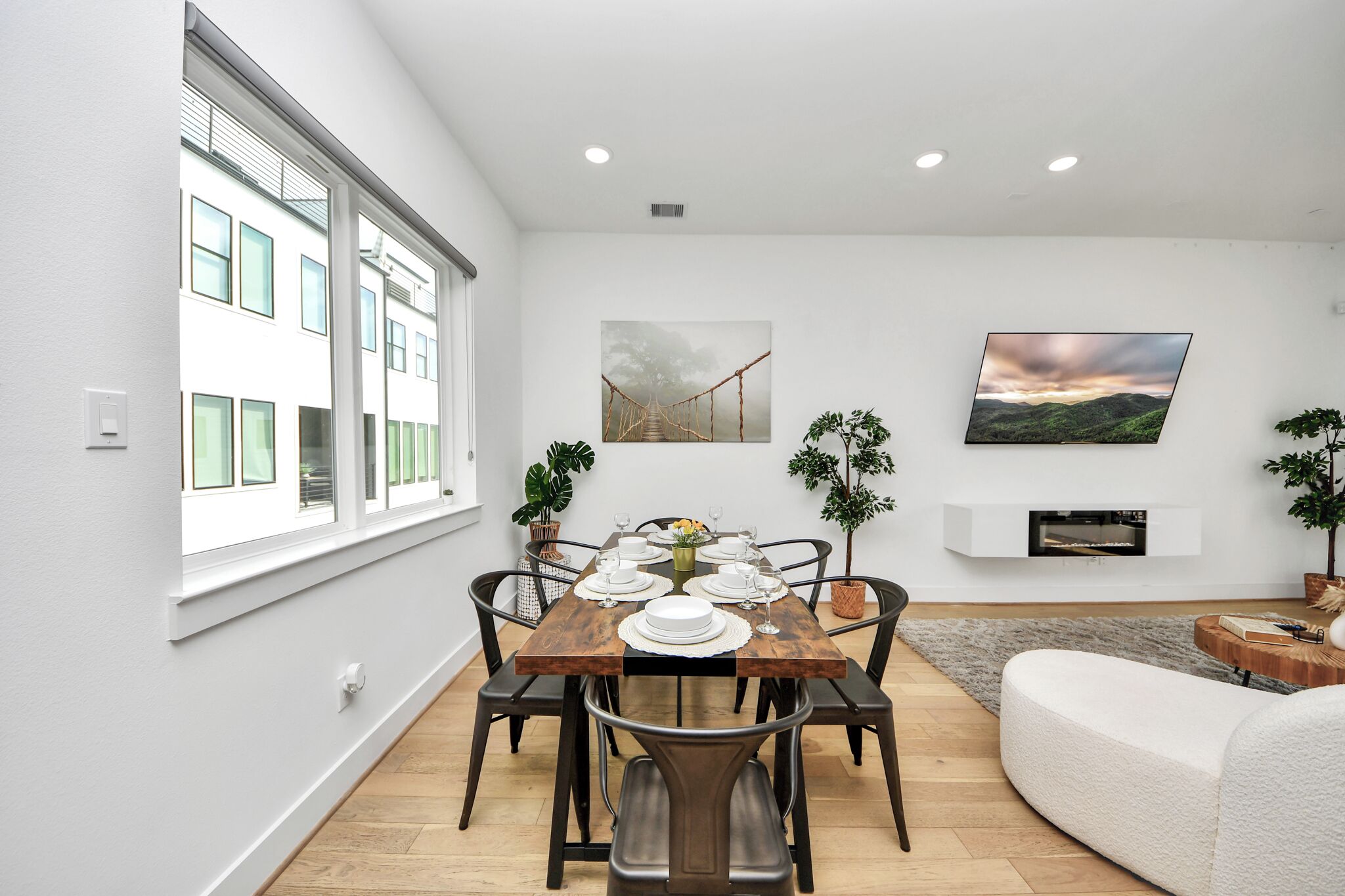 Un coin repas avec une table en bois dressée pour six personnes, entourée de chaises, avec un canapé blanc et une cheminée en arrière-plan.