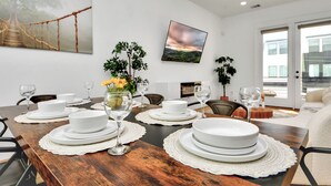 Uma sala de jantar com uma mesa de madeira posta para refeição, pratos brancos e copos. Há duas plantas em vasos, um quadro na parede e vista de um edifício através de uma porta de vidro.
