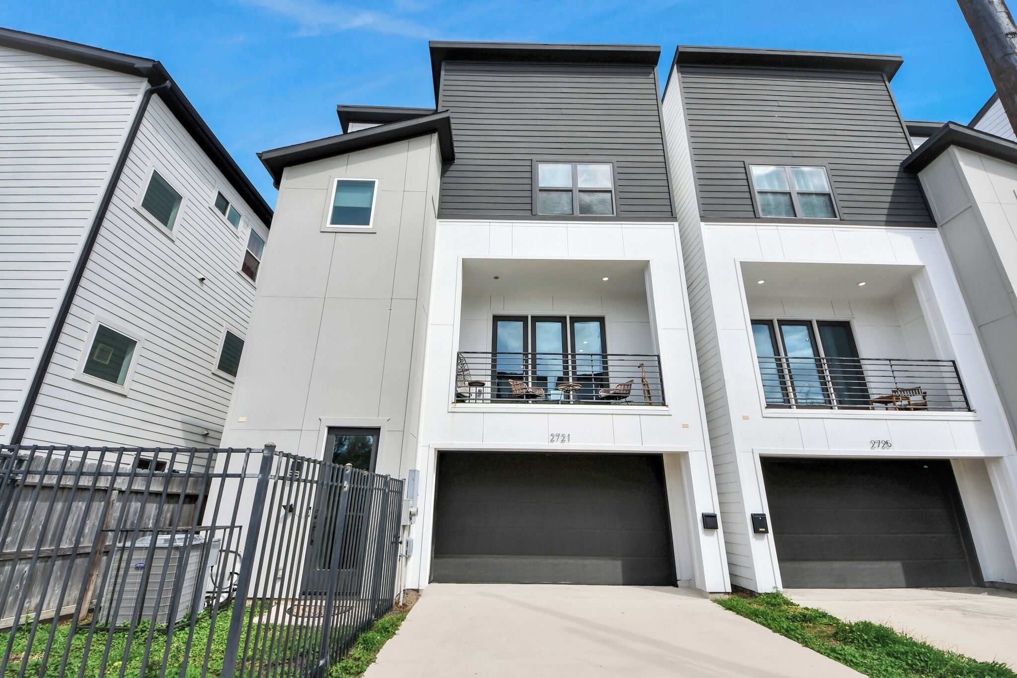 A modern multi-story residential building with two garage doors, a balcony with outdoor seating, and a visible address number.