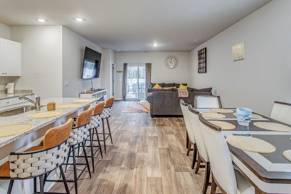A modern kitchen with a long counter and bar stools, a dining area with a table and chairs, and a living room with a sofa and TV.