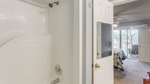 A bathroom with a bathtub, a shower, and a door leading to a bedroom with a bed and a chair.