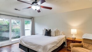 Un dormitorio con cama, ventilador de techo, mesita de noche, lámpara, silla y vistas a través de una puerta corrediza de vidrio.