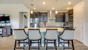 A modern kitchen with dark cabinets, a central island with bar stools, and a TV in the background. - 2 Mi to TPC San Antonio: Group Getaway w/ Theater! (San Antonio)