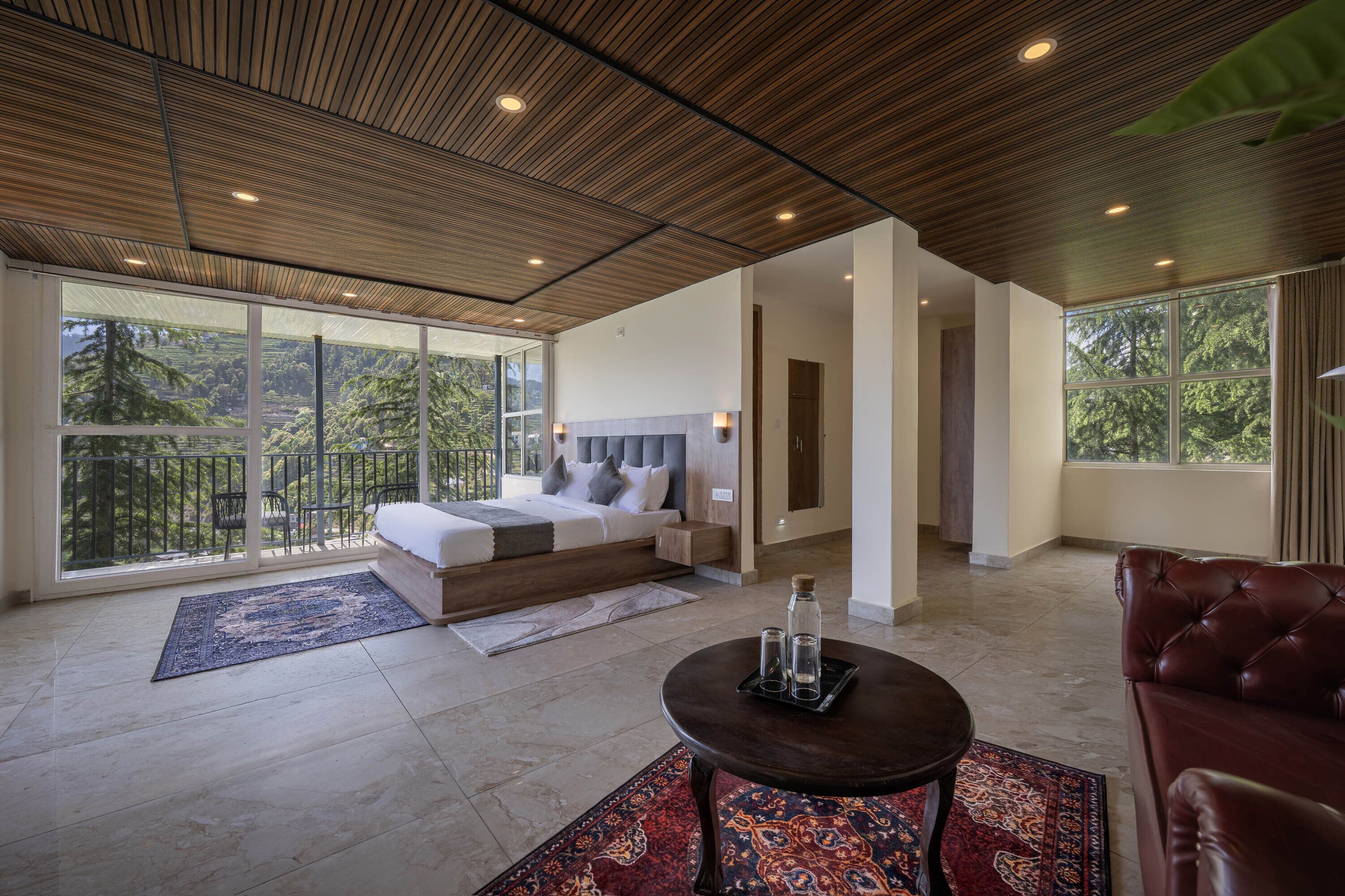 A modern bedroom with a large bed, a sitting area with a coffee table, and a balcony with a view of trees.