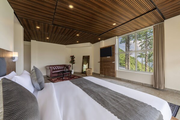 A spacious bedroom with a large bed, a wooden ceiling, a TV, and a large window with a view of trees. - Echor Alpine Meadows Khajjiar Dalhousie (Chamba)