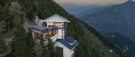 A modern two-story mountain house with a balcony, surrounded by dense forest and mountain views.