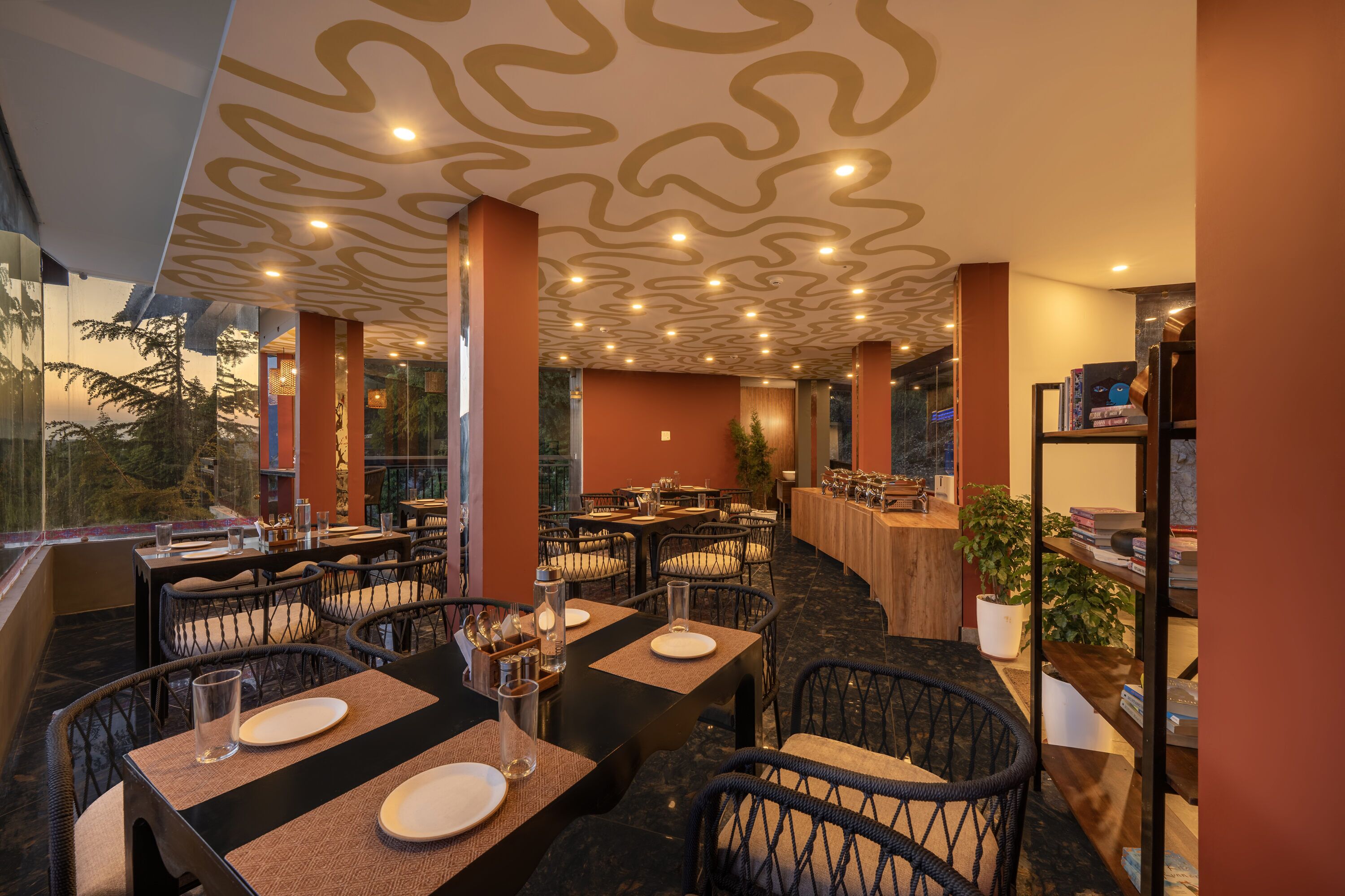 Un restaurant avec des tables dressées pour le service, un plafond décoré et une vue sur les arbres à l’extérieur.
