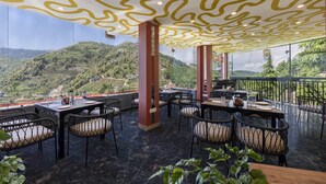An outdoor dining area with tables and chairs, a view of a green landscape, and a ceiling with a wavy pattern. - Echor Alpine Meadows Khajjiar Dalhousie (Chamba)