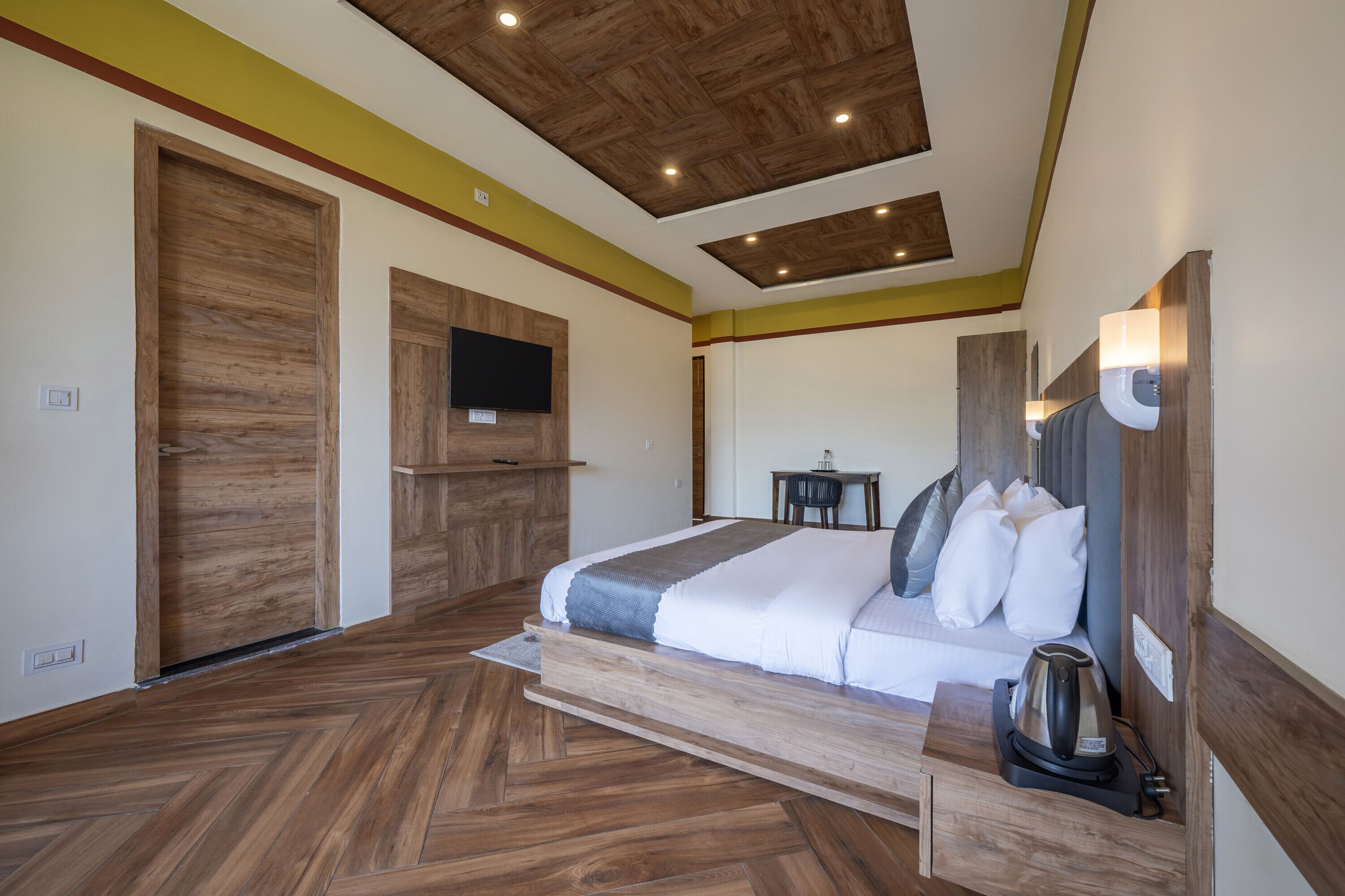 A modern hotel room with a wooden floor, a bed with white bedding, a flat-screen TV, a small table with a kettle, and a wooden ceiling.