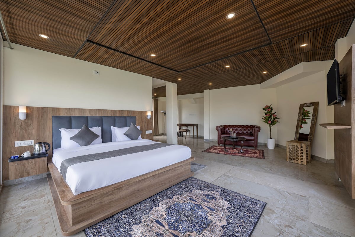 Un dormitorio moderno con cabecera de madera, cama grande, mesita de noche, cafetera, televisor, sofá rojo, planta en maceta y espejo.