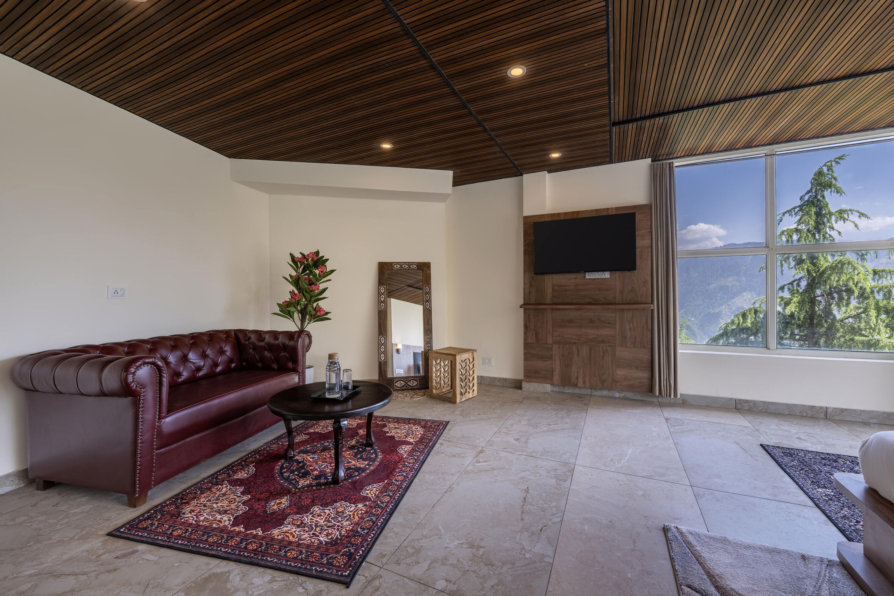 A living room with a leather sofa, a coffee table, a patterned rug, a wooden ceiling, a flat-screen TV, and a large window with a mountain view.