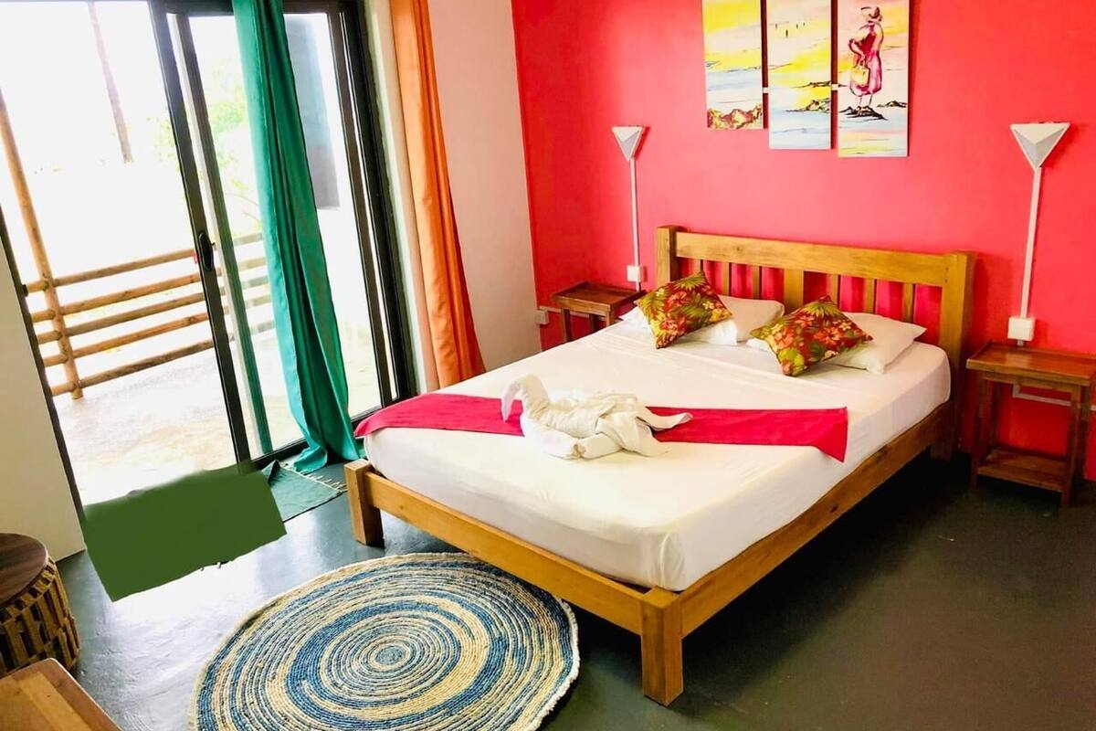 A hotel room with a bed, a green exit door, a view of the outside, and a painting on the wall.