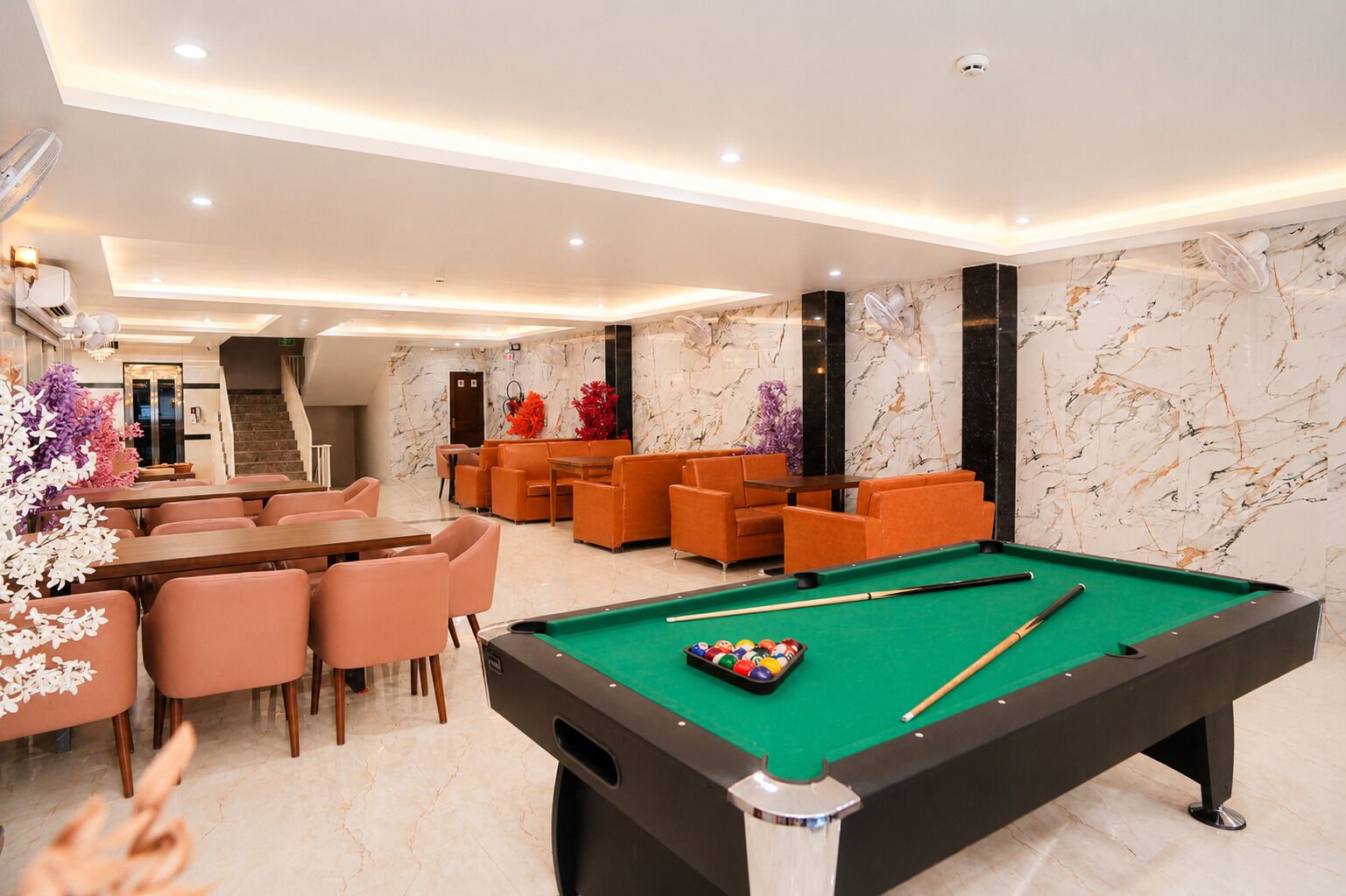 A pool table with a green felt surface, surrounded by orange chairs and a dining table with pink chairs.