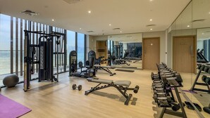 Une salle de sport équipée de divers appareils d’exercice, dotée de grandes fenêtres et offrant une vue sur la ville.