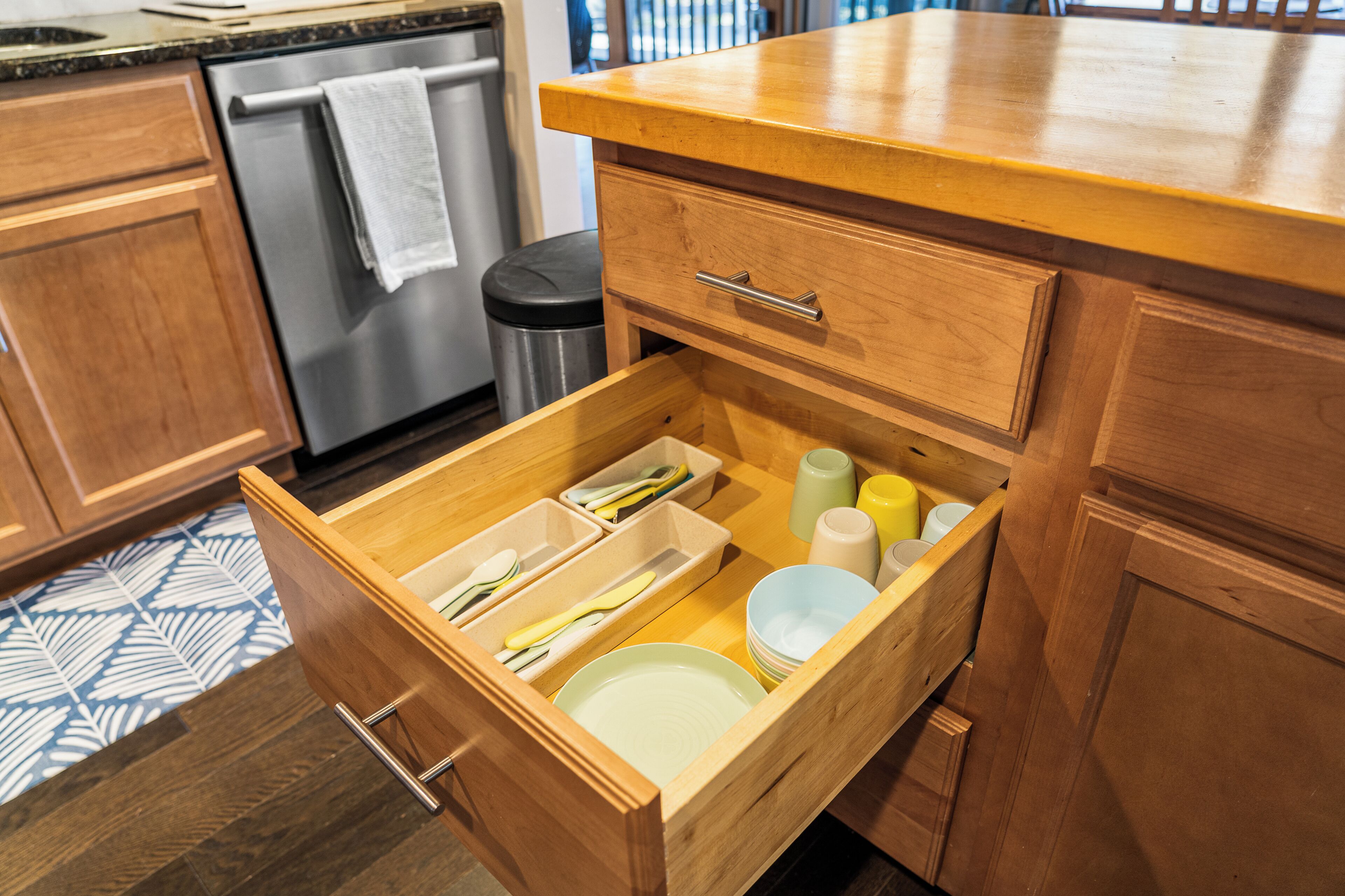 Cocina con armarios de madera, lavavajillas de acero inoxidable y un organizador de cajones con utensilios y platos.