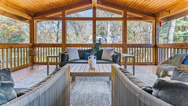 Eine überdachte Veranda mit Holzbalken, einer Couch, einem Couchtisch und Blick auf Bäume.