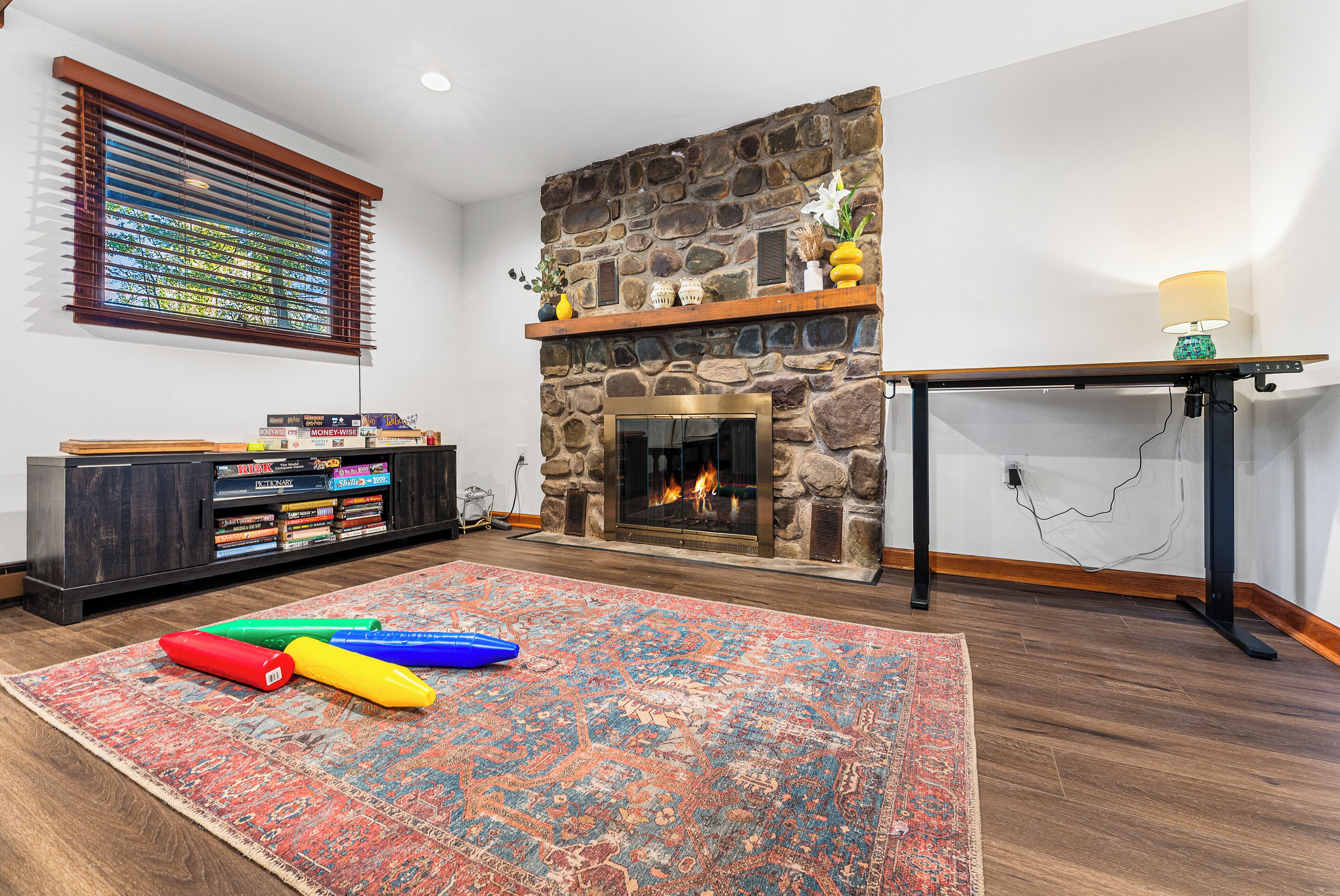 A living room with a stone fireplace, a bookshelf, a colorful rug, and a window with blinds.