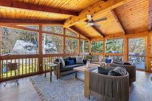 Um deck de madeira com sofá e cadeiras, um ventilador de teto e grandes janelas.
