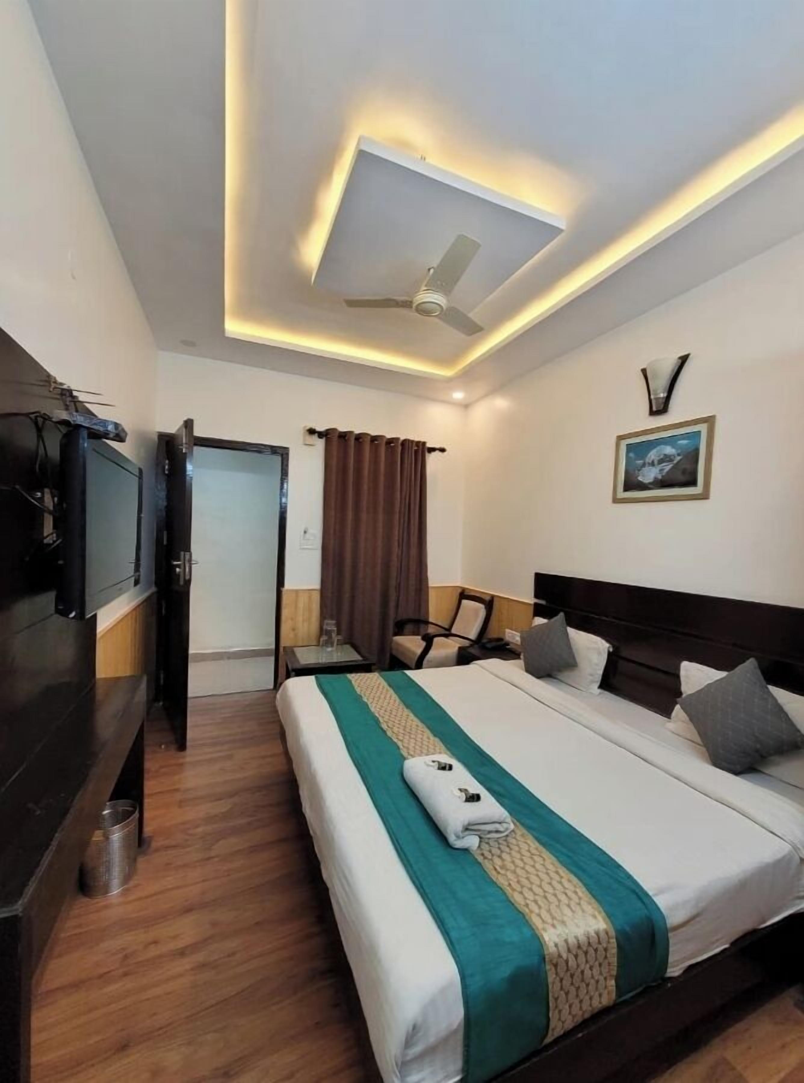 A hotel room with a bed, a TV, a ceiling fan, a painting, and a door.