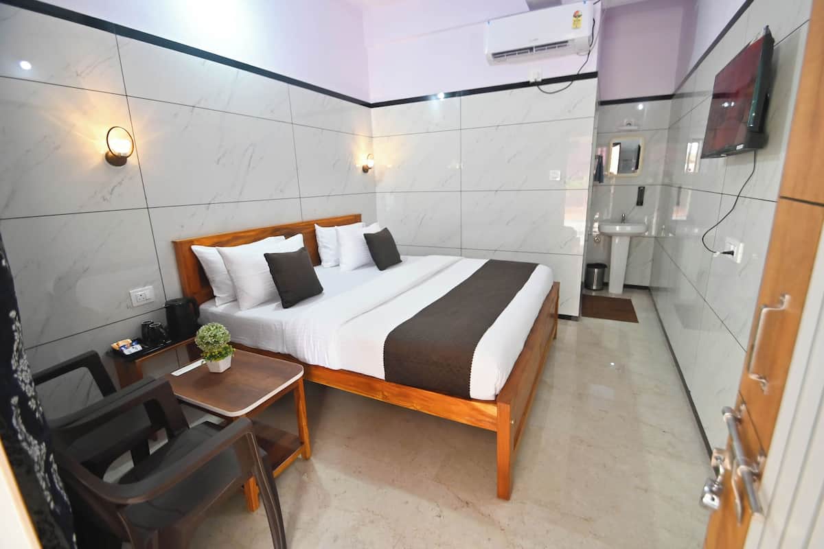 Quarto de hotel com cama, mesa de cabeceira, cadeira, planta em vaso pequeno e televisão fixada na parede.
