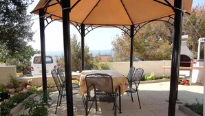 Eine überdachte Terrasse mit Tisch und Stühlen, umgeben von Pflanzen und Blick auf die Straße.